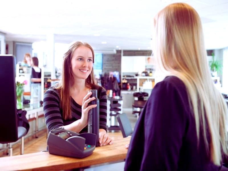 A smiling salon employee hands a frizz-free haircare product to a blonde customer at the checkout counter in a modern hair salon.