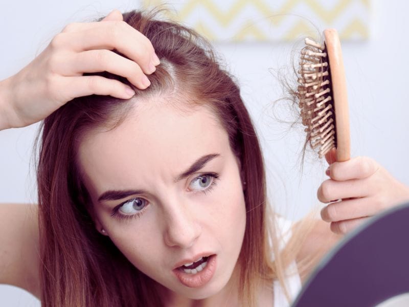 Worried woman examines thinning hair and hair loss on brush, looking into mirror in concern.