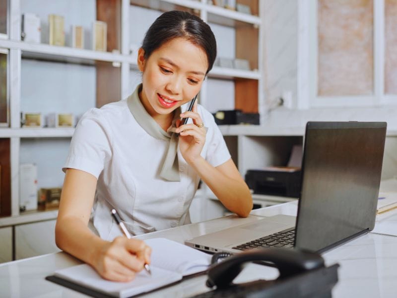 Salon receptionist on phone and writing in a notebook, managing client bookings and appointments.