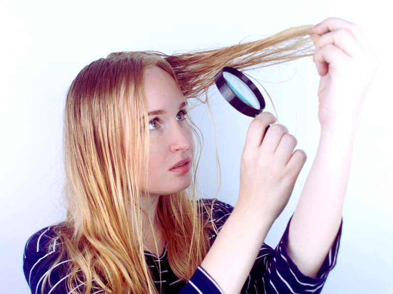 Woman inspects oily, flat hair strands with a magnifying glass, showing concern on her face.