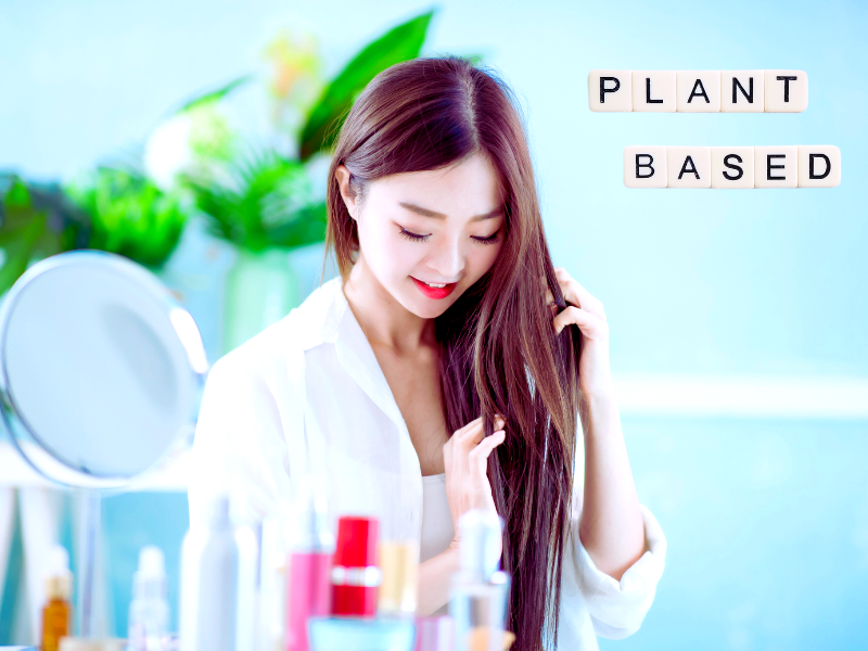 Smiling woman admires her healthy hair at vanity with text “PLANT BASED” in scrabble tiles.