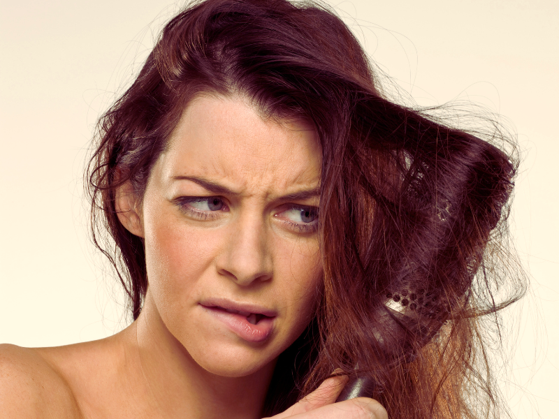 Frustrated woman with tangled, damaged hair caught in a round brush—signs of poor hair care habits.