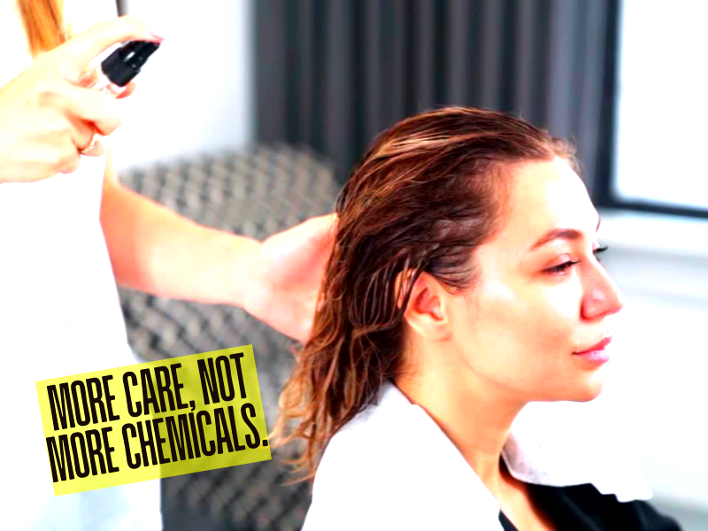 Hair stylist spraying treatment on wet hair with bold text: "More care, not more chemicals."