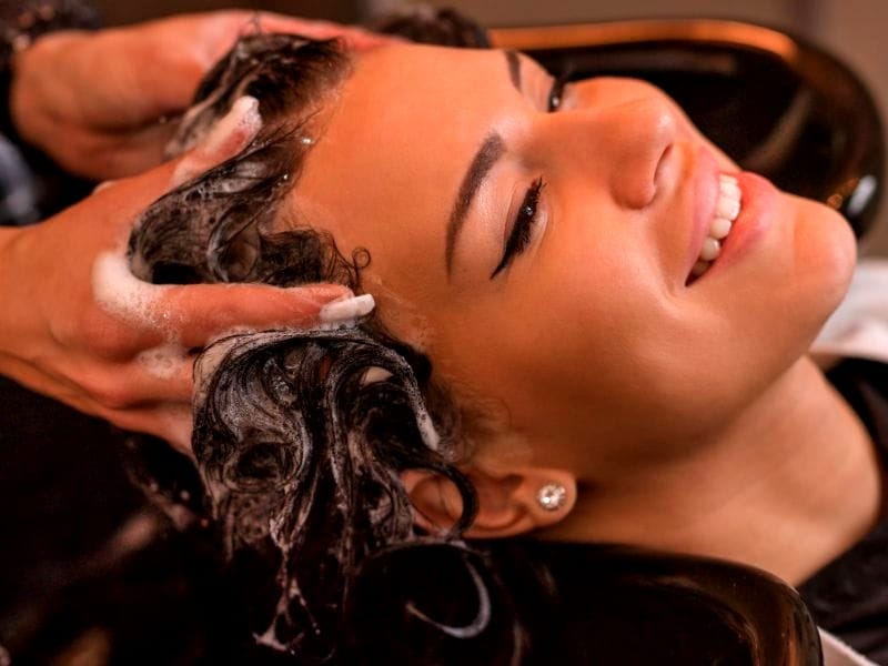 Smiling woman enjoying a relaxing scalp massage and shampoo at a hair salon sink with foam lather.