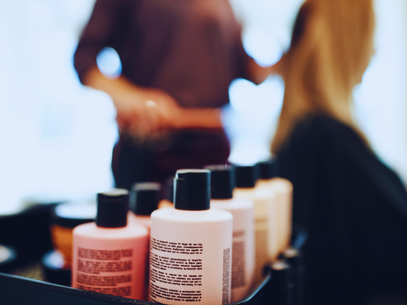 Haircare bottles lined up on a salon trolley with a hairstylist working on a client in the blurred background.